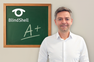 Jan Šimík, ředitel BlindShellu, stojí sebevědomě v bílé košili vedle tabule s logem BlindShell a velkým nápisem A+. Z celého obrázku vyzařuje silný pocit pozitivity.