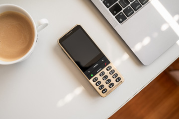Pískově zlatý telefon BlindShell Classic 3 je položen na stole mezi hrníčkem s kávou a laptopem.