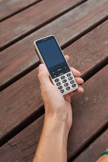 Pískově zlatý telefon BlindShell Classic 3 v lidské dlani. V pozadí jsou vidět tmavě hnědá prkna.