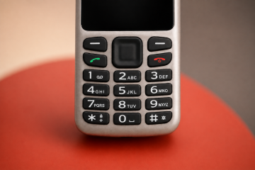 Detail klávesnice telefonu BlindShell Classic Lite na jemně rozostřeném pozadí v teplých béžových, šedých a červených tónech. Snímek zdůrazňuje velká, dobře hmatatelná číselná tlačítka.