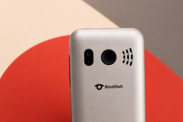 Detail horní části zadní strany stříbrného telefonu BlindShell Classic Lite na geometrickém pozadí v teplých béžových, červených a tmavě šedých tónech. Snímek zdůrazňuje kulaté SOS tlačítko a logo BlindShell.