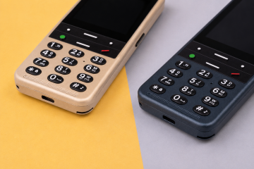 Detail dvou telefonů BlindShell Classic 3 v barvách Zlatavě pískové a Oceánově modré, umístěných vedle sebe na jemně žlutém a světle šedém pozadí. Snímek zdůrazňuje jejich velké hmatové klávesnice s dobře rozlišitelnými tlačítky, tlačítka pro ovládání hovorů, mřížky reproduktorů a nabíjecí konektory.