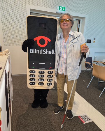 Jeden z výherců telefonu BlindShell Classic 3 hrdě pózuje vedle nového maskota BlindShellu. Se slunečními brýlemi, lehkou bundou a bílou holí stojí usměvavě vedle maskota. Společně zachycují hřejivý okamžik ze SightCity 2026 plný radosti, vděčnosti a pocitu komunity, který dělá podobné akce tak výjimečnými.