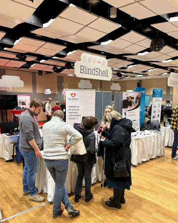 Návštěvníci se shromažďují u stánku BlindShell na konferenci INSPO zaměřené na asistivní technologie. Poslouchají produktové ukázky, ptají se na dotazy a živě diskutují v rušném prostředí výstavní haly.