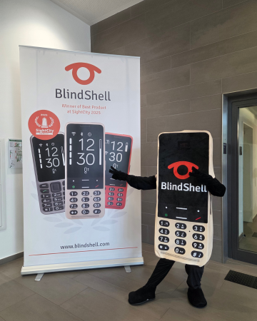 Náš nový maskot BlindShellu, navržený jako obří zlatý telefon BlindShell s klávesnicí, displejem a výrazným logem BlindShell, hrdě pózuje vedle reklamního banneru BlindShell. Díky svému hravému vystupování a přátelské energii se maskot rychle stal oblíbencem návštěvníků, rozdával úsměvy a přinesl na SightCity 2026 ještě více zábavy.