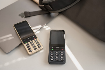 The image shows two BlindShell Classic 3 phones placed on a table next to a laptop and a backpack. The colors of the phones are golden sand and ocean blue, both featuring large tactile keypads. The phones are angled slightly, with their screens turned off.