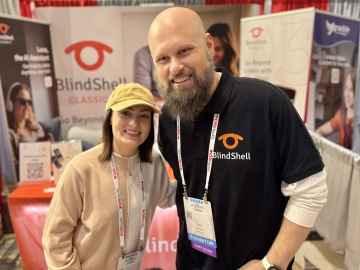 Molly Burke a Sam z The Blind Life stojí blízko sebe a usmívají se u stánku BlindShell během konference o asistivních technologiích. Molly má na sobě světlou kšiltovku, béžový svetr a konferenční visačku na šňůrce. Sam má černé polo tričko BlindShell s logem a vystavovatelskou visačku. Oba působí uvolněně a přátelsky. V pozadí jsou vidět bannery a propagační materiály BlindShell.
