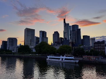 Panorama Frankfurtu při západu slunce s moderními mrakodrapy odrážejícími poslední paprsky dne. Charakteristické výškové budovy se tyčí nad řekou Mohan, podél jejíhož nábřeží kotví lodě. Růžové a oranžové mraky se rozprostírají po večerní obloze a vytvářejí malebný pohled na finanční metropoli Německa, často přezdívanou „Mainhattan“.