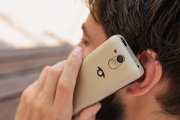 Muž drží telefon BlindShell Classic 3 v barvě pískově zlaté u ucha během telefonního hovoru. Fotografie zachycuje zadní stranu telefonu s fotoaparátem, SOS tlačítkem a logem BlindShell.