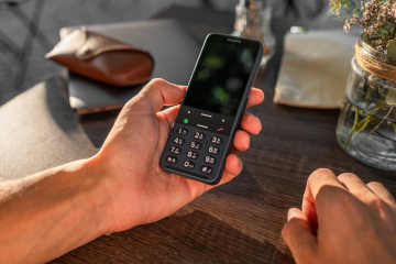 Muž drží telefon BlindShell Classic 3 v barvě Oceanově modré v jedné ruce při sezení u venkovního stolku. Na fotografii je dobře vidět velká hmatová klávesnice a jednoduché rozložení tlačítek navržené pro snadné ovládání nevidomými a slabozrakými uživateli.