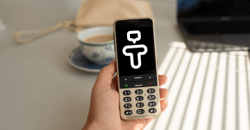 Ruka držící telefon BlindShell Classic 3 v barvě Pískově zlaté s logem Tandem na displeji. Za telefonem je bílo-modrý hrnek. Celý obrázek je plný kontrastních barev.